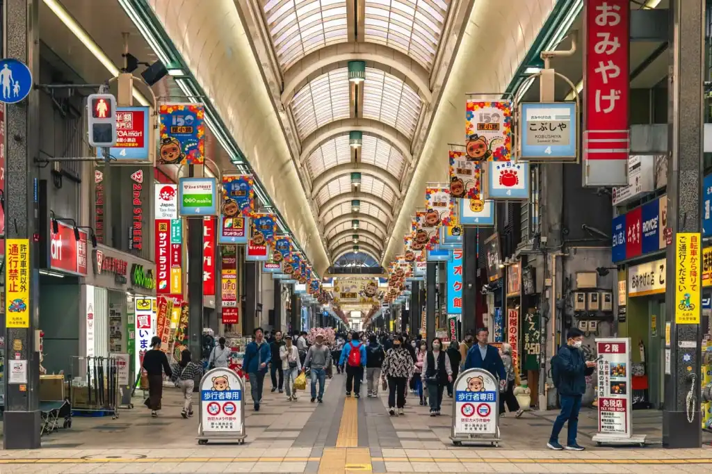 Shoppers walking through a bustling covered Japanese shopping arcade with vibrant signs, colorful banners, and diverse retail stores, capturing the lively atmosphere of Tokyo shopping districts.