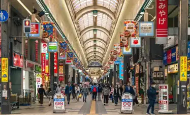 Shoppers walking through a bustling covered Japanese shopping arcade with vibrant signs, colorful banners, and diverse retail stores, capturing the lively atmosphere of Tokyo shopping districts.