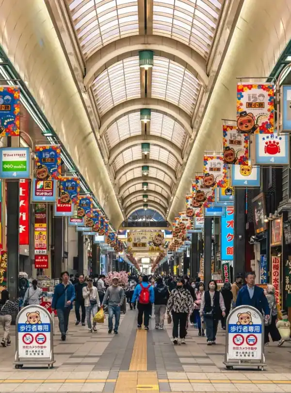 Shoppers walking through a bustling covered Japanese shopping arcade with vibrant signs, colorful banners, and diverse retail stores, capturing the lively atmosphere of Tokyo shopping districts.