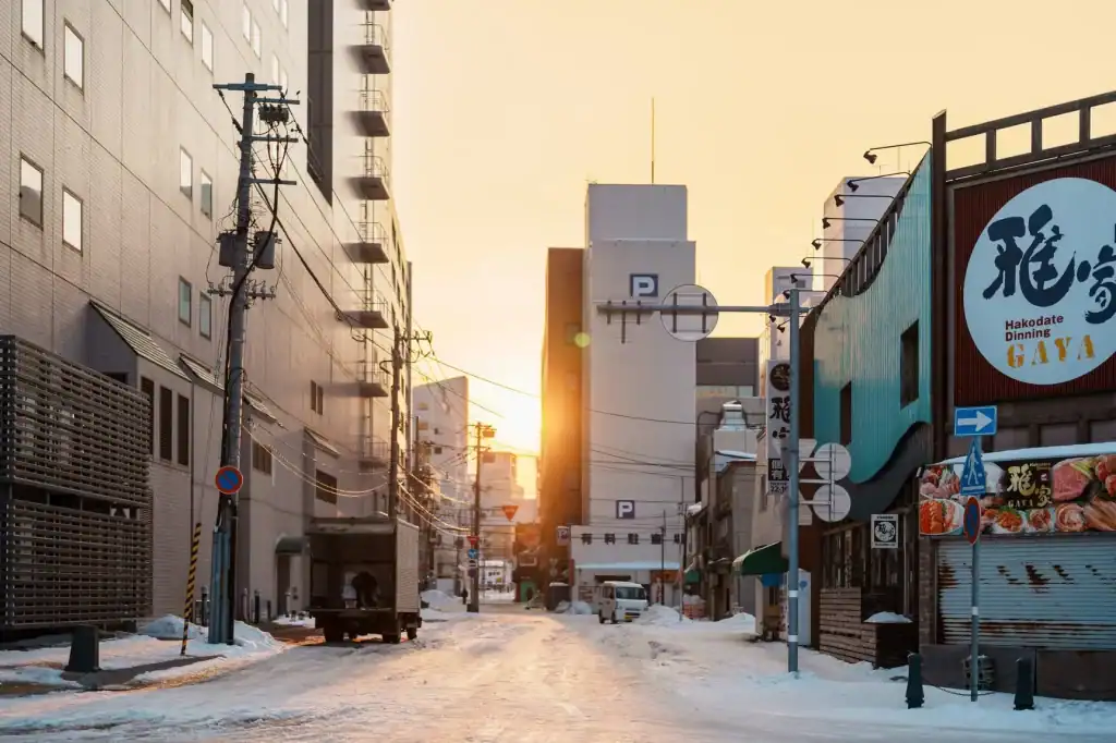 Serene winter street scene in Hakodate, Japan during sunset, showcasing snow-covered roads, urban buildings, and traditional dining signage for an authentic travel experience.