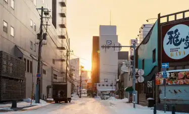 Serene winter street scene in Hakodate, Japan during sunset, showcasing snow-covered roads, urban buildings, and traditional dining signage for an authentic travel experience.