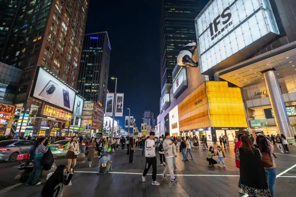 Vibrant city street scene at night with illuminated billboards, busy pedestrians, and high-rise buildings showcasing luxury brands and popular stores, capturing the energetic urban nightlife.