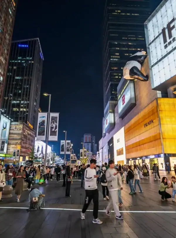 Vibrant city street scene at night with illuminated billboards, busy pedestrians, and high-rise buildings showcasing luxury brands and popular stores, capturing the energetic urban nightlife.